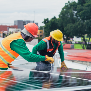 Two workers in safety gear installing solar panels on a rooftop. One worker is adjusting a panel while the other monitors the process. Buildings and trees are visible in the background.